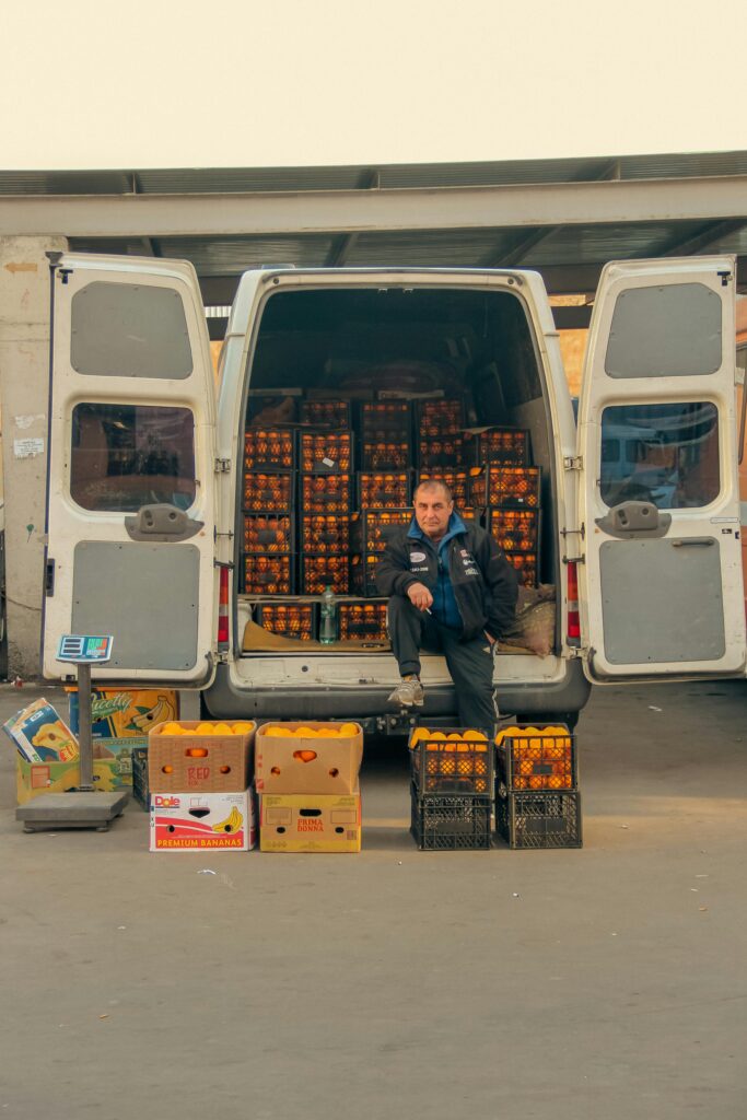 pexels-photo-19036286-19036286 A man selling fruit from a van in an urban market setting, surrounded by boxes of produce.